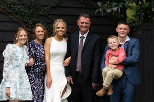 A group of six people are posing together outdoors. There are four adults and two children. One adult is wearing a white dress, another suit