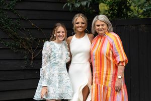 Three women are posing together outdoors. The woman on the left is wearing a light blue floral dress, the woman in the middle is in a white