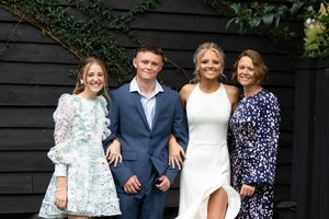 A group of four people posing in front of dark wooden wall with greenery. The first person on the left is wearing a light blue floral dress,