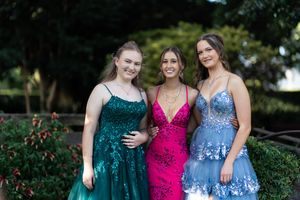 Three people are standing together outdoors, dressed in formal gowns. The person on  left is wearing a green dress, the person in the middle