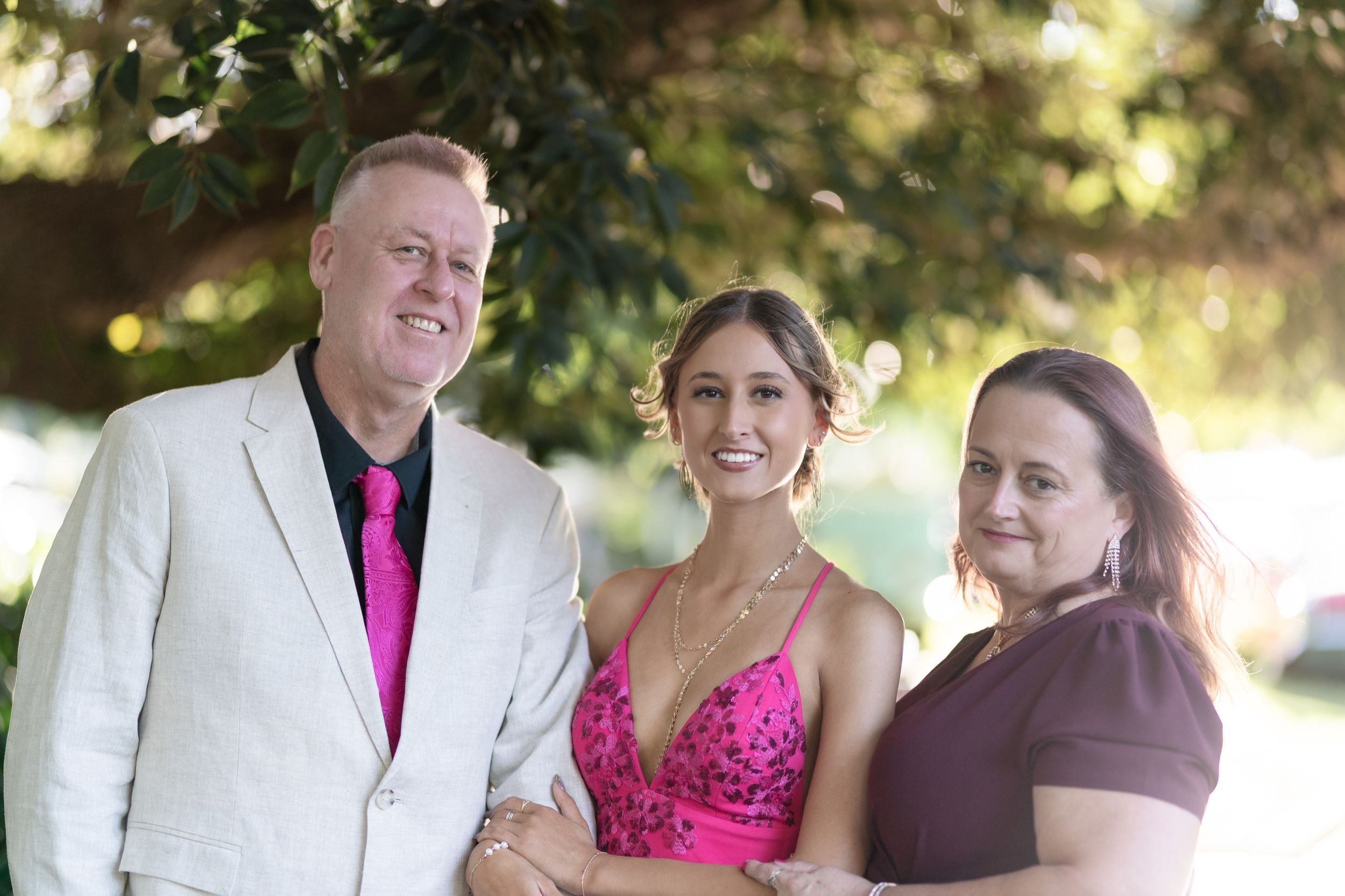 A group of three people posing outdoors The person on the left  wearing a light-colored suit with a bright pink tie The person in the middle