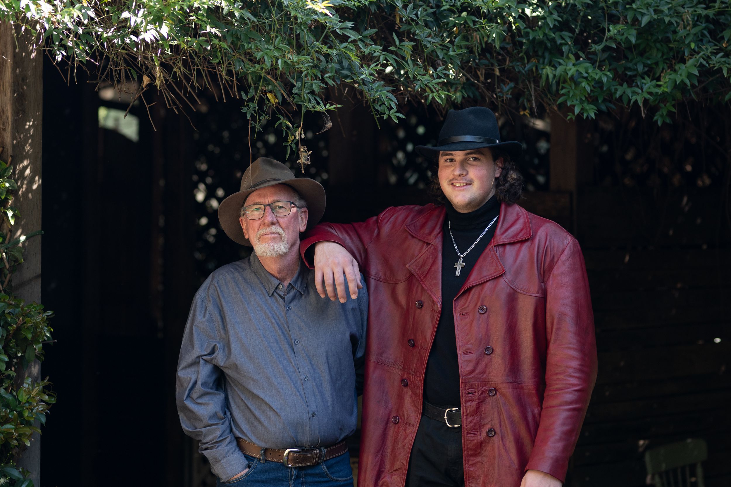 Two people are standing together outdoors. The person on the left is wearing a gray shirt and a brown hat, while the person on the right