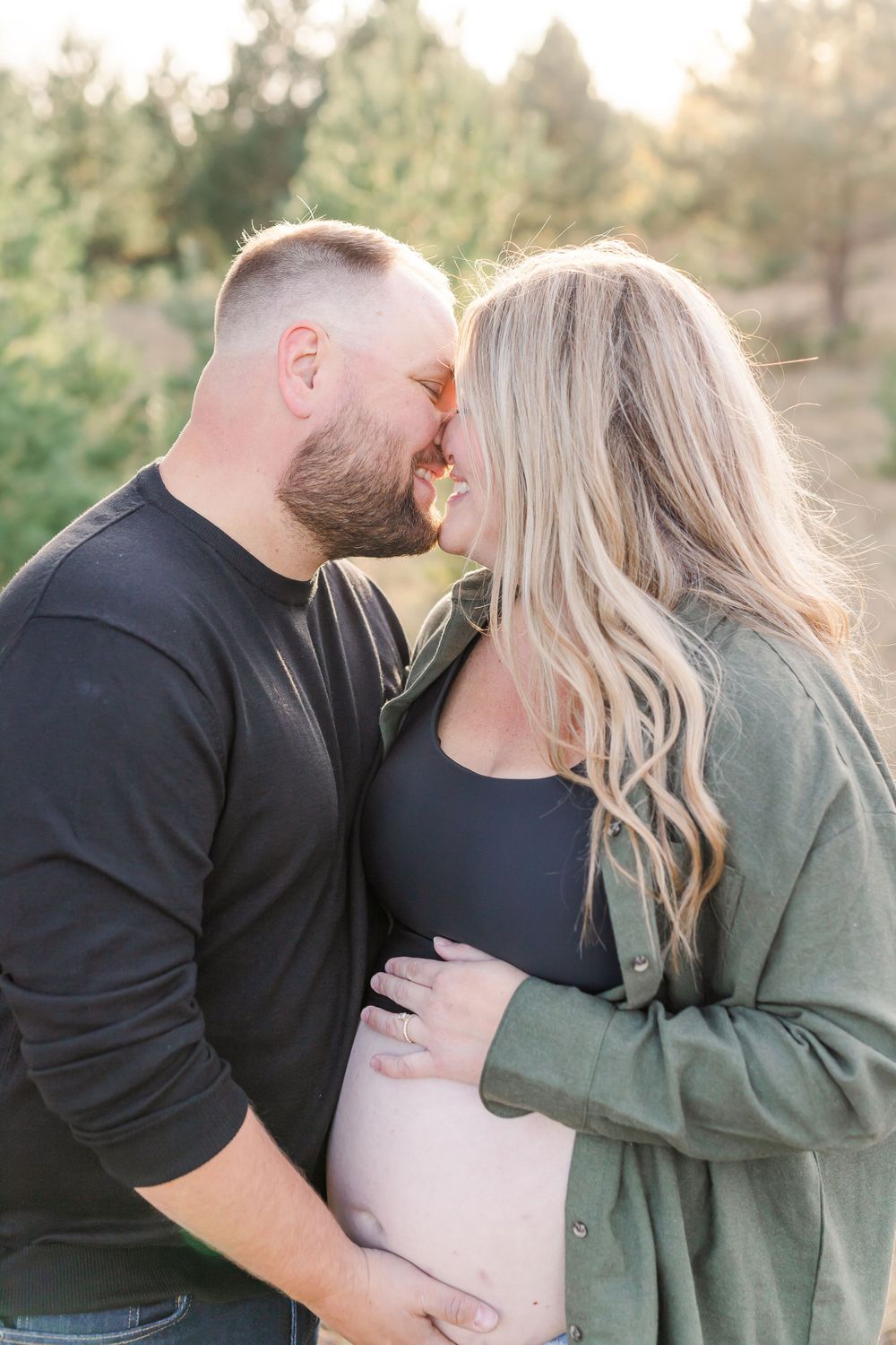 Intimate maternity portrait outdoors with couple embracing and touching foreheads in soft sunlight.