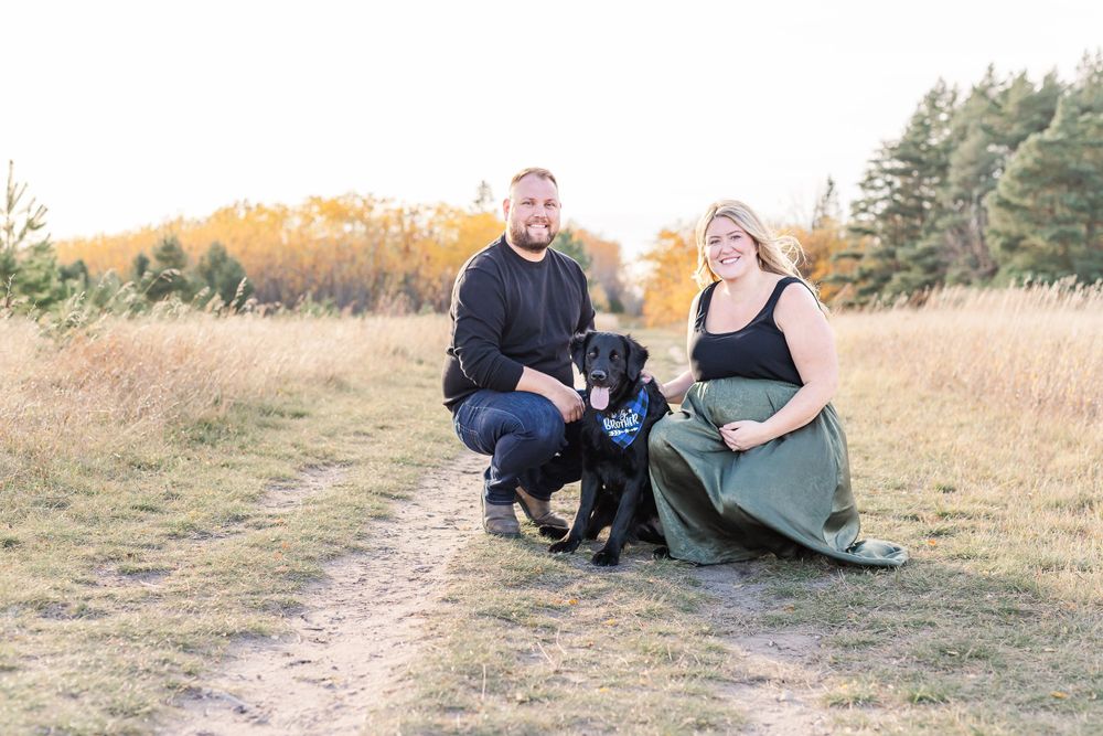 A couple and their black dog pose together on a dirt path in an autumn field at sunset.