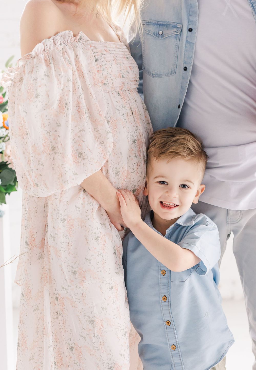 A young child wearing a blue button-up shirt stands next to someone in a flowy floral off-shoulder dress.