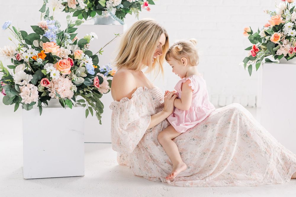 A parent and child wearing light-colored dresses share a tender moment beside floral arrangements on a white background.