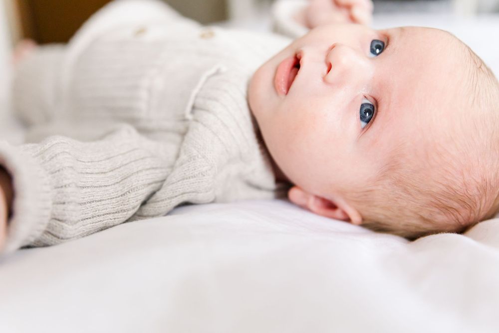 A smiling baby in a white sweater lies on soft bedding looking up with bright blue eyes.