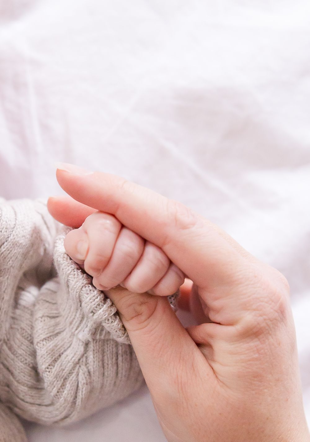 A small baby's hand gently holding an adult's finger while wrapped in a soft gray knit blanket.