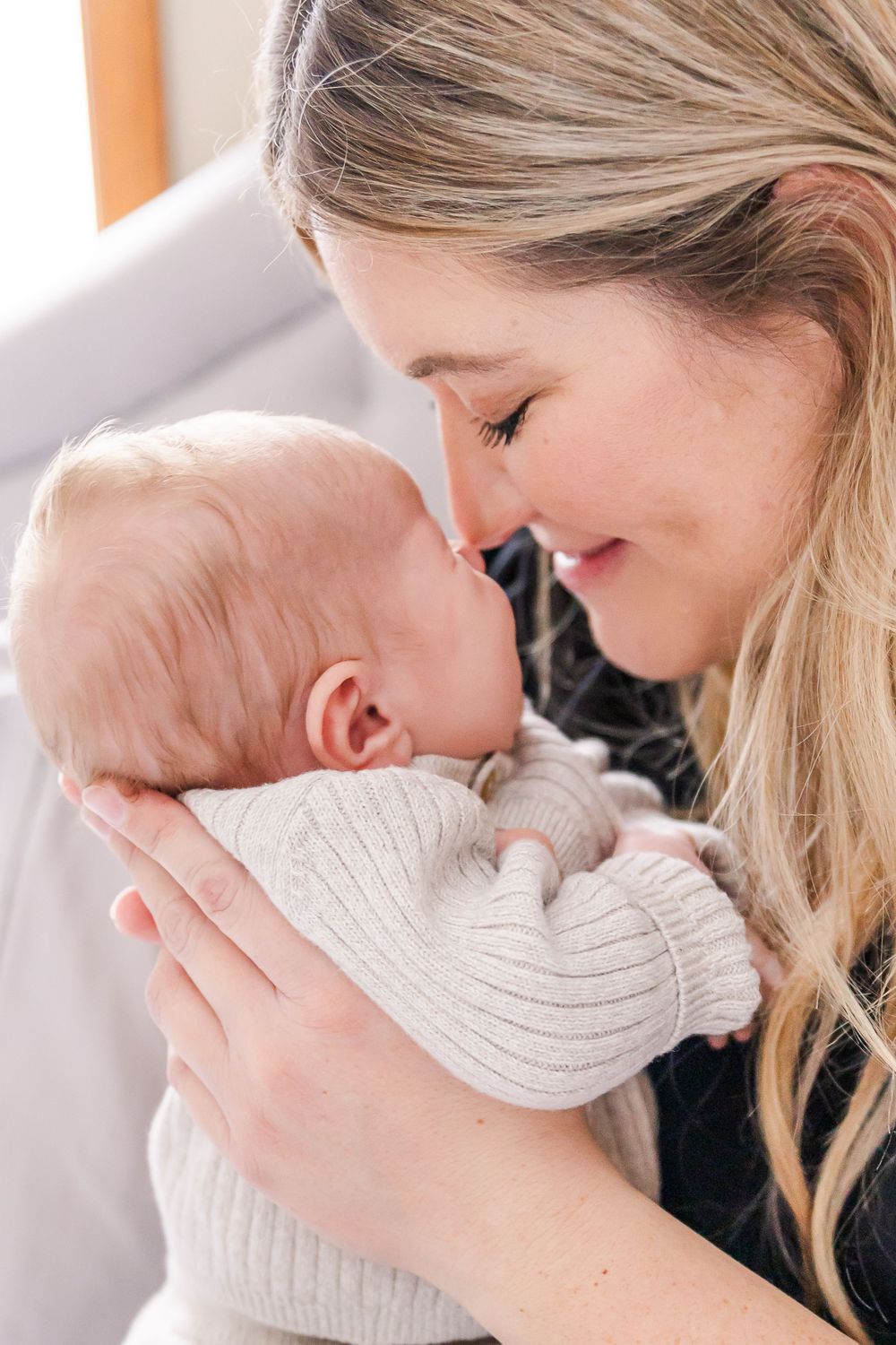 A tender moment as mother in black cradles baby wearing white knit sweater in loving embrace.