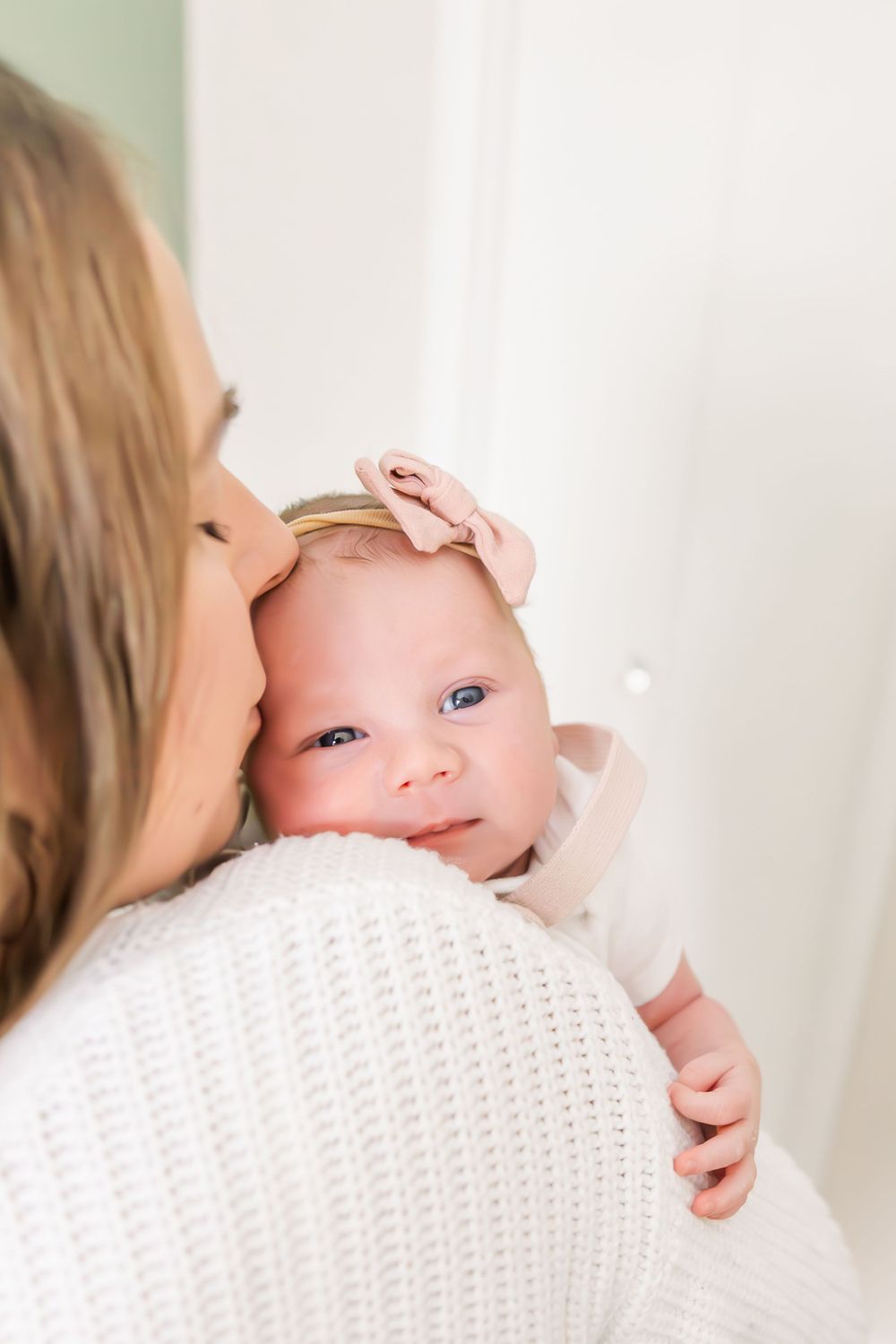Baby wearing pink headband peeks over shoulder while being held in white knit sweater.