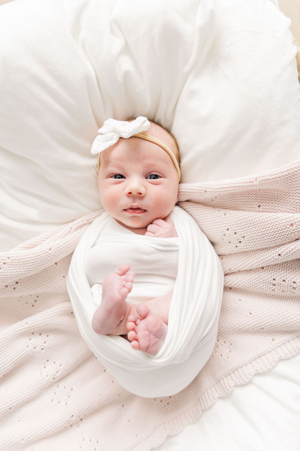 Baby wrapped in soft white swaddle blanket lying on cream knit blanket with white bow headband.