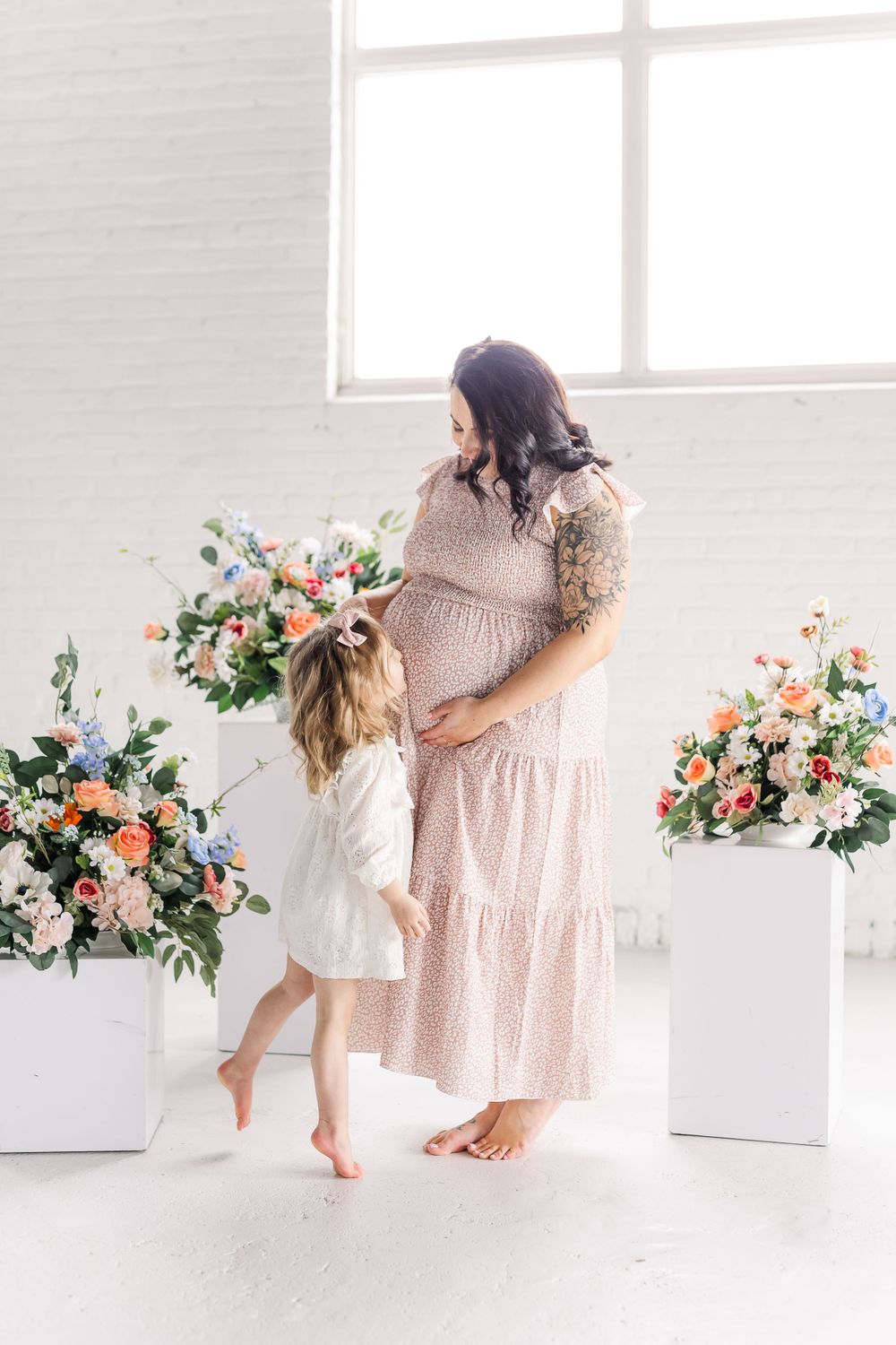 Mother and daughter share a tender moment in white room with soft pink floral arrangements and natural light.