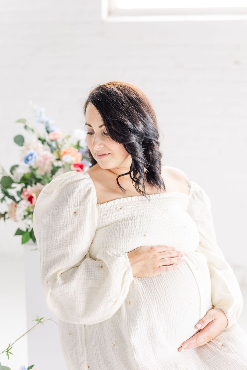 Expectant mother in flowing white dress and sweater poses gracefully against floral backdrop in bright airy setting.