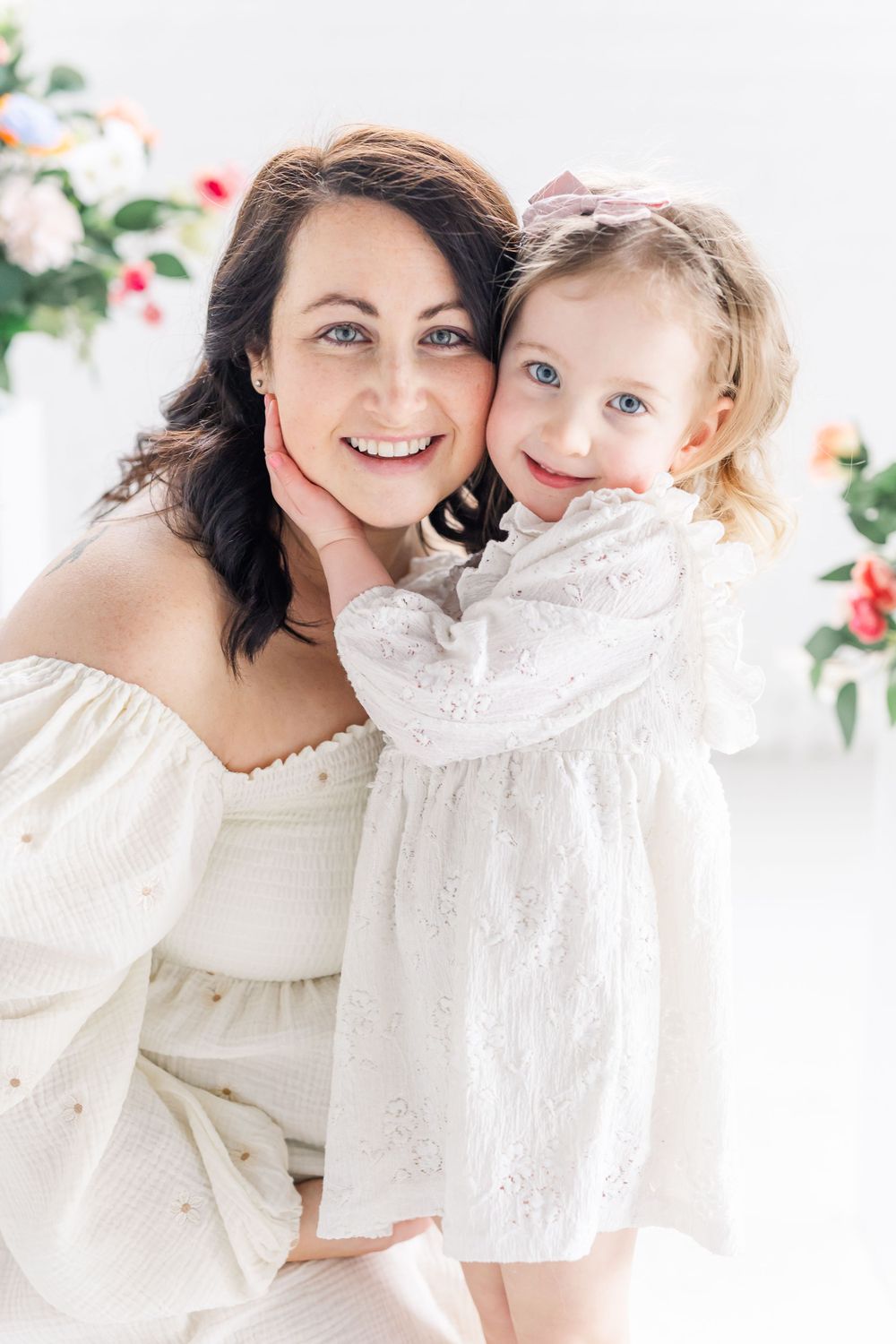 A sweet portrait of a mother and daughter in matching white lace dresses posing against a floral backdrop.