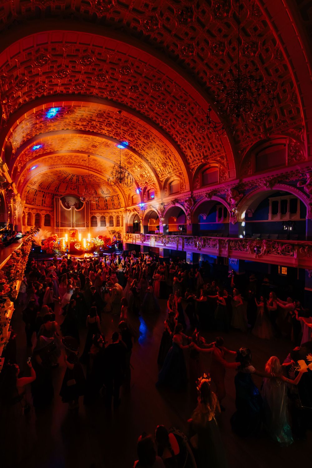 Elegant ballroom with vaulted ceiling filled with guests under dramatic red and blue theatrical lighting.