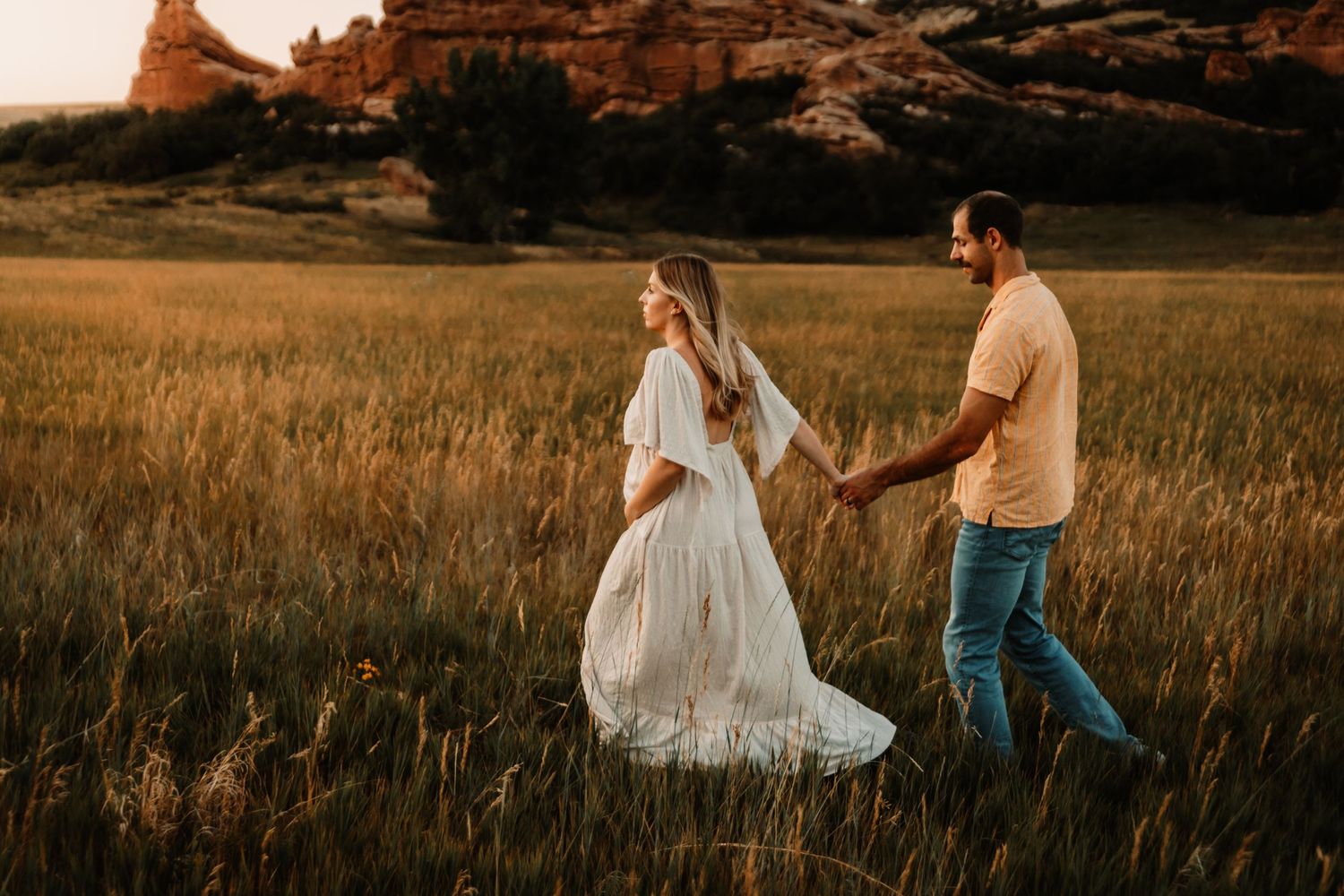A couple walks hand in hand through a golden field at sunset with red rock formations in the background.