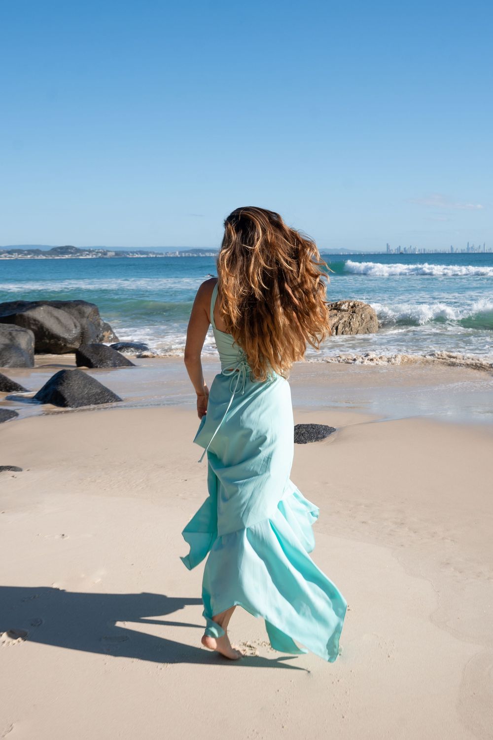 Woman in flowing teal maxi dress walking barefoot on sandy beach with ocean waves and rocks in background.