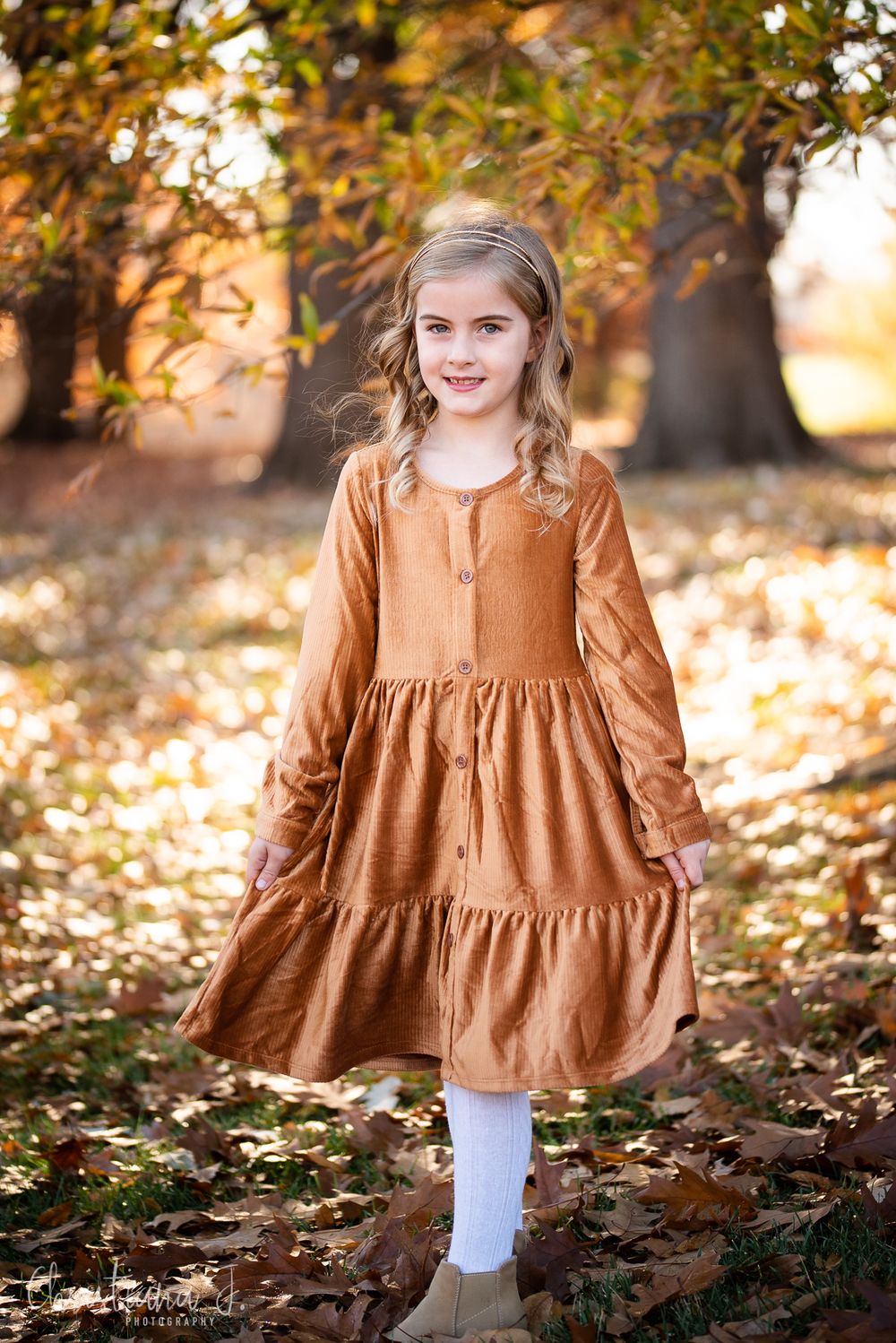 Young girl in rust-colored tiered dress poses among autumn leaves with golden trees in background.