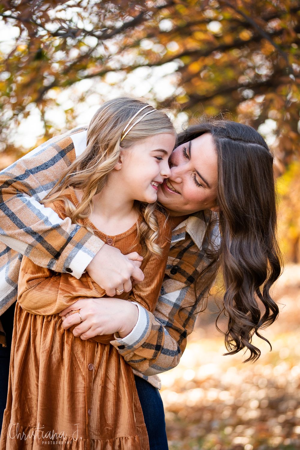 A mother and daughter share a tender nose-to-nose moment outdoors, wearing plaid and rust tones amid fall foliage.