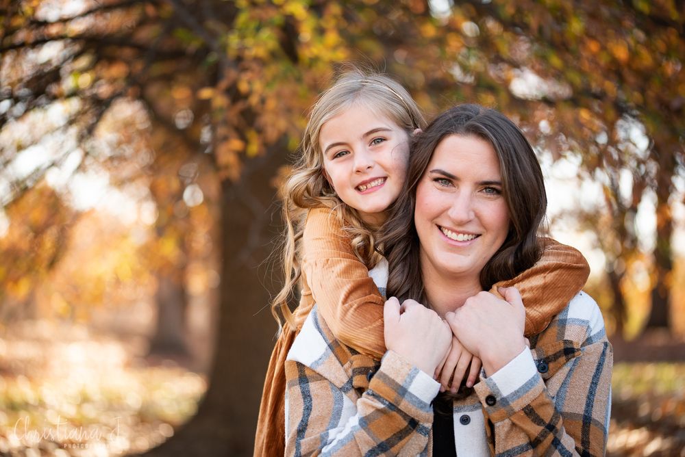 A smiling mother and daughter pose together outdoors among autumn trees, the girl hugging her mom from behind at Loose Park in Kansas City