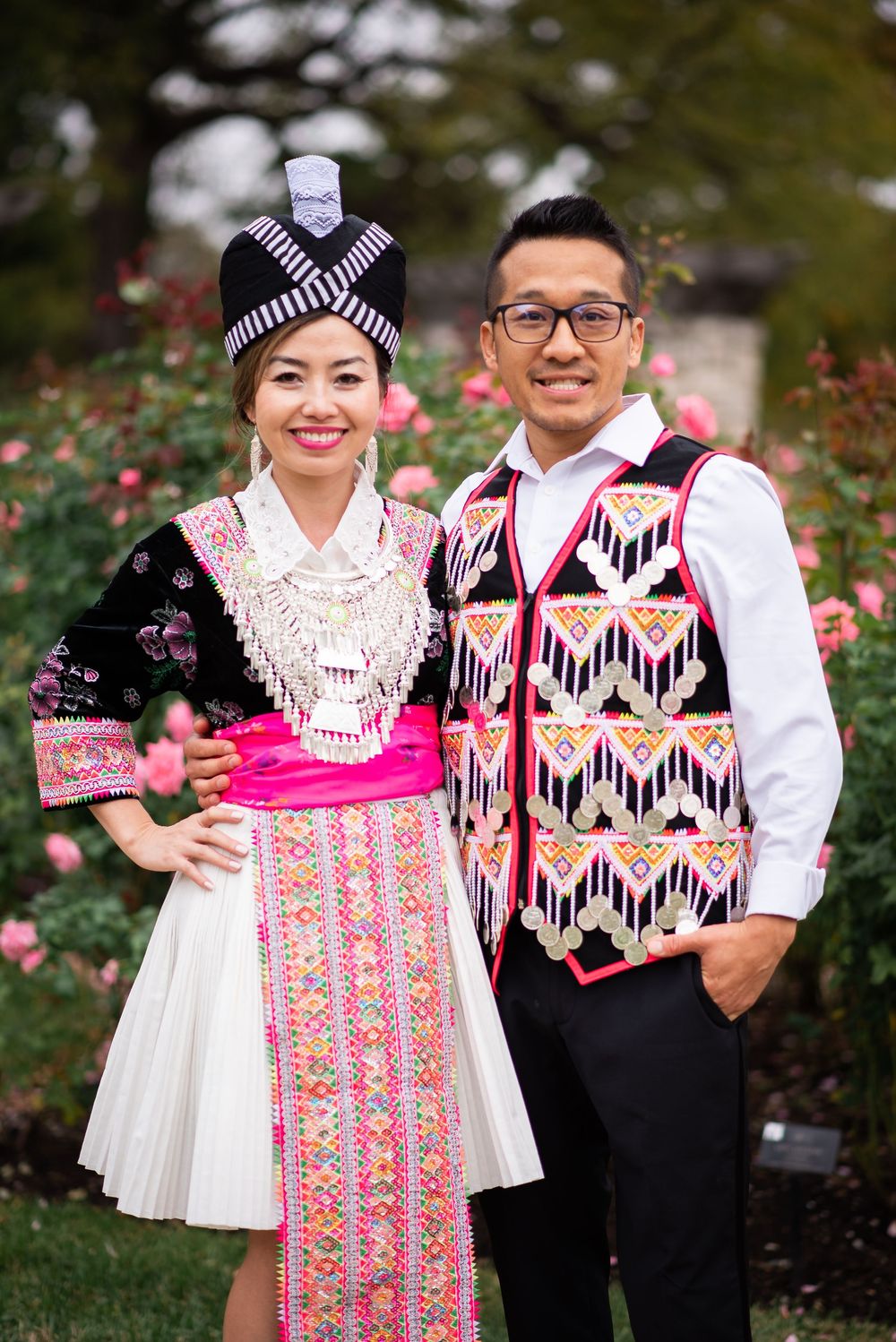 Couple wearing traditional Hmong attire with colorful embroidery and beadwork, posing in front of blooming roses.