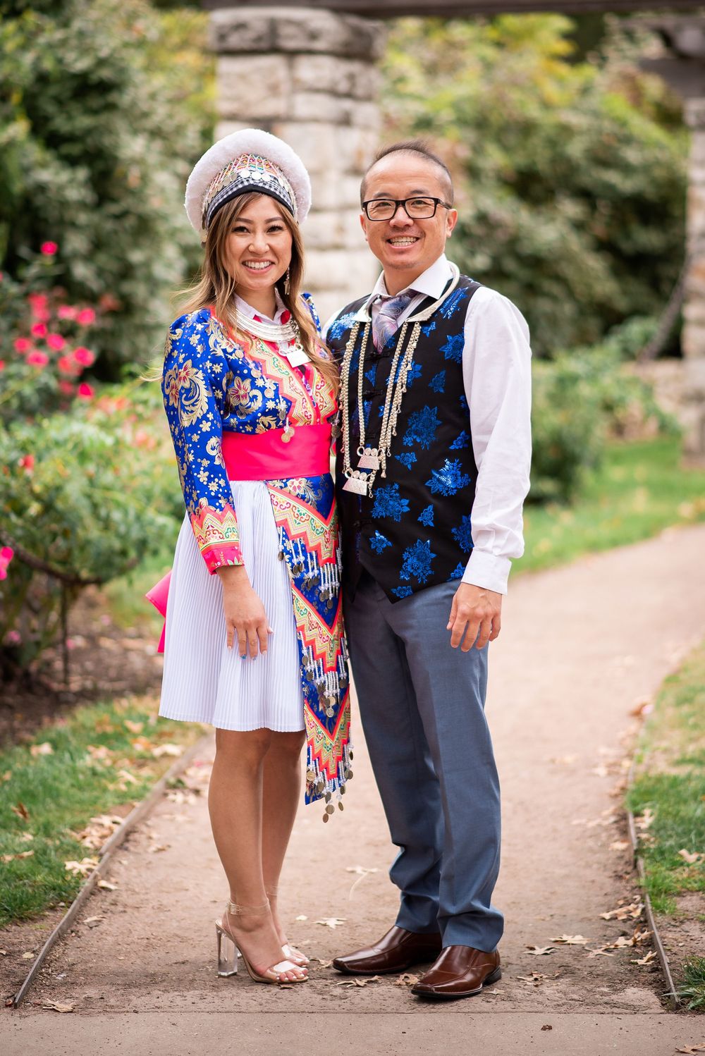 Couple posing in traditional Hmong attire in a garden, woman in colorful embroidered dress with white hat, man in floral vest.