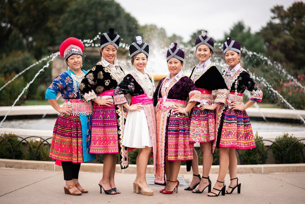 Six women in vibrant traditional Hmong attire pose together near a fountain in the Rose Garden at Loose Park kansas city
