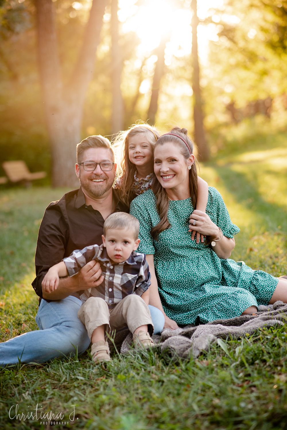 Family of four sitting on a blanket in a sunlit park, smiling warmly during a golden hour outdoor photo session.