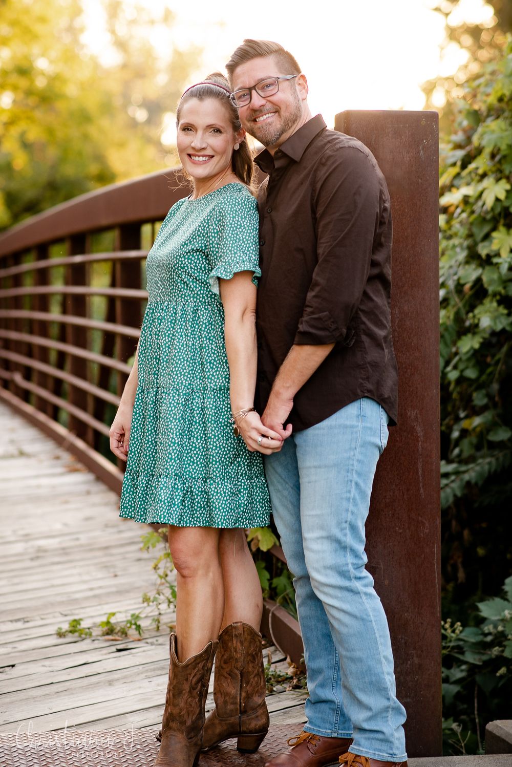 A smiling couple poses on a wooden bridge, woman in teal floral dress with cowboy boots, man in dark shirt and jeans.
