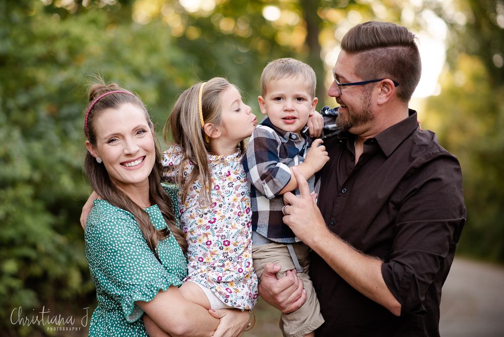 A happy family of four poses outdoors, with parents holding two young children sharing a sweet kiss in Leawood, Kansas