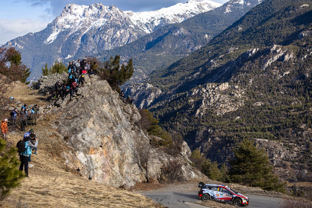 Rally car racing on mountain road with snow-capped peaks in background.