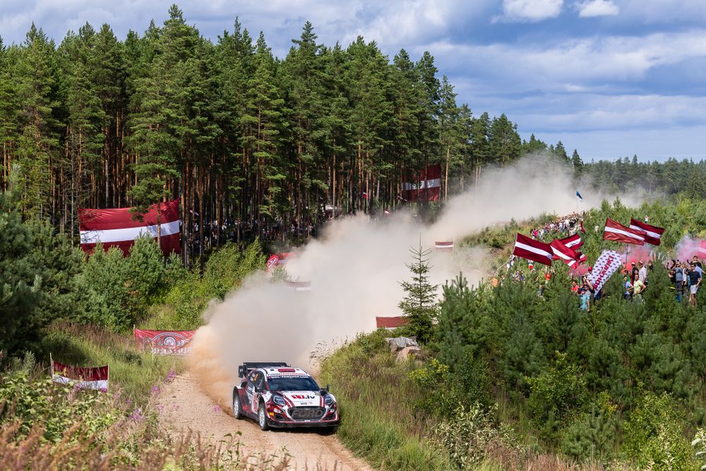 Rally car kicking up dust on a forest track with spectators waving flags along the course.