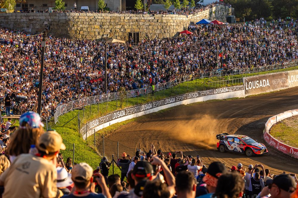 Large crowd of rally spectators watching a racing car navigate a dirt track curve at sunset.