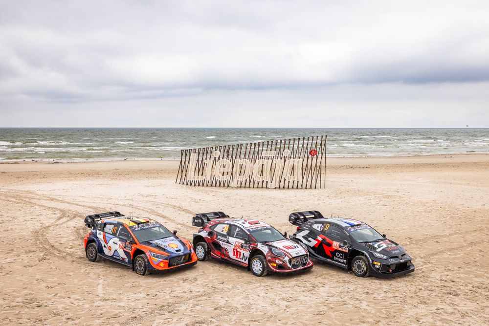 Three rally cars parked on a sandy beach with a wooden fence and dramatic cloudy sky in the background.
