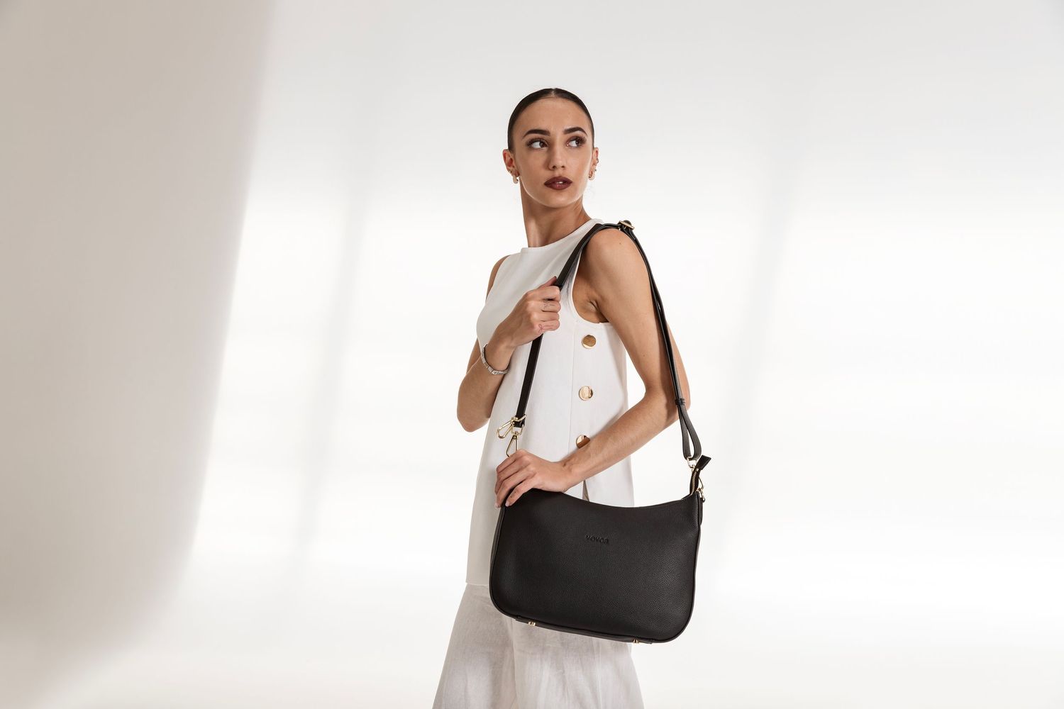 Woman in white sleeveless dress holding a black leather shoulder bag against a light background.