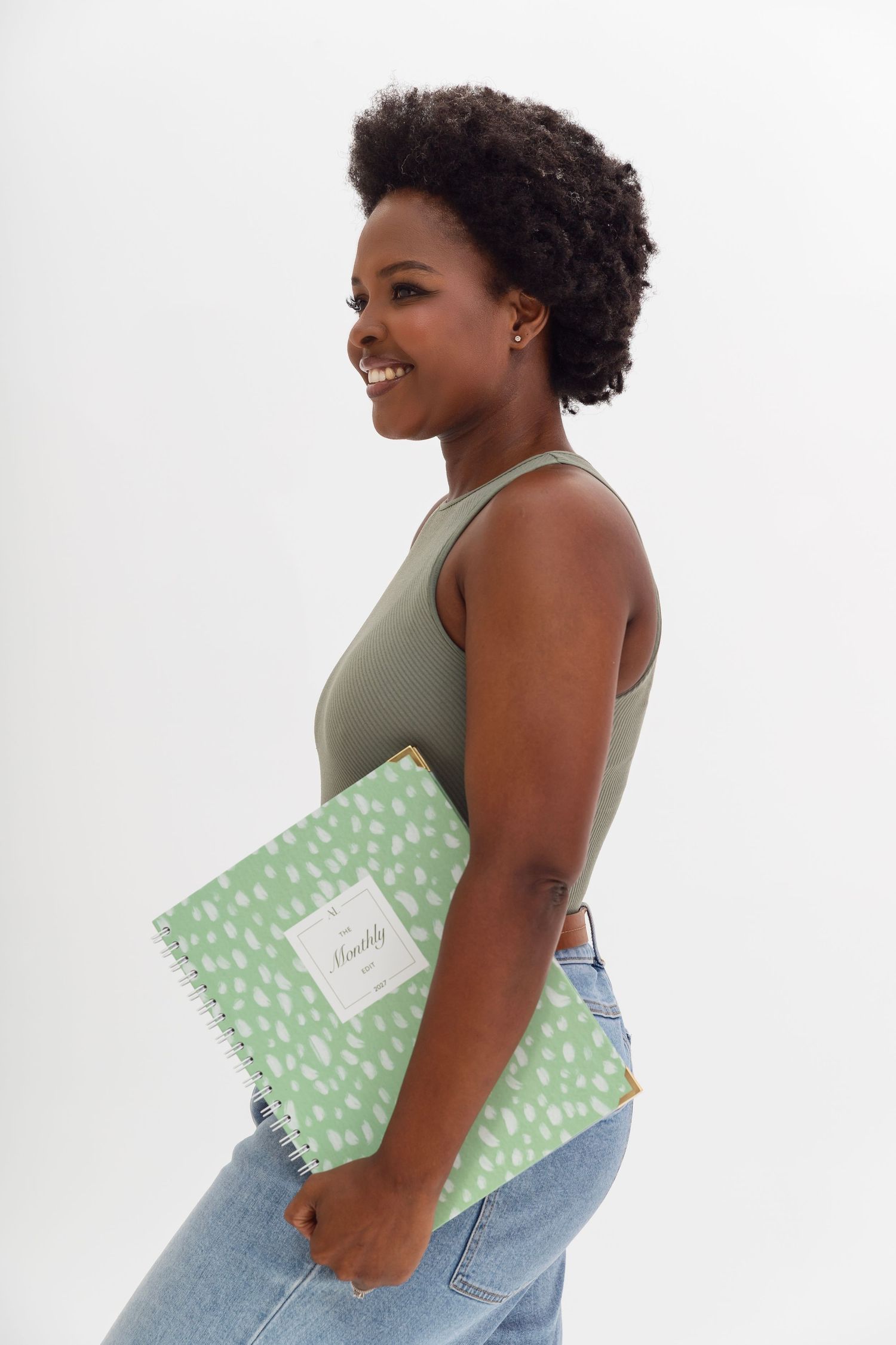 Smiling young Black woman holding a green spiral notebook, wearing a sage tank top and jeans on white background.