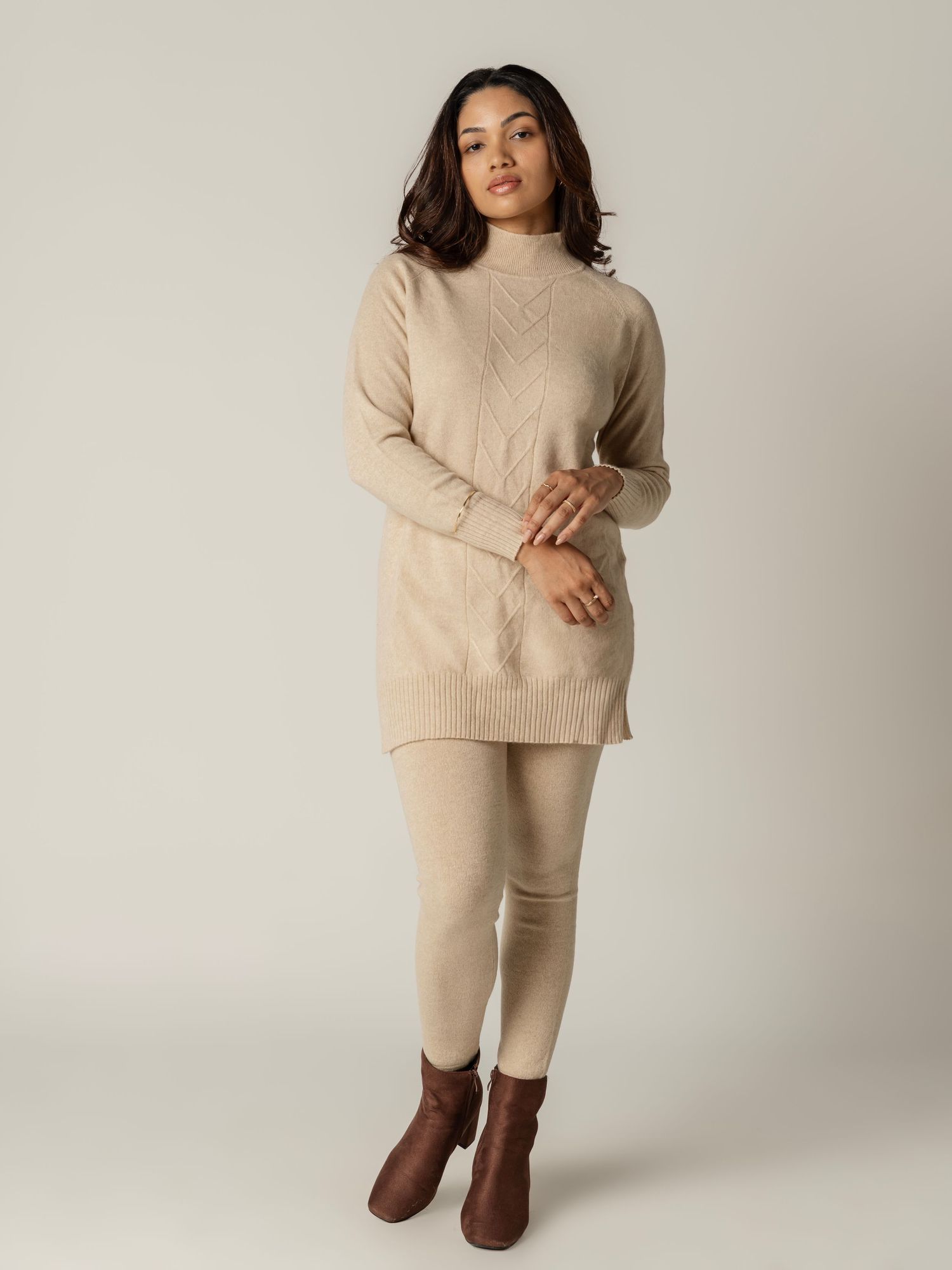 Model wearing a beige knit turtleneck sweater with matching leggings and brown ankle boots against neutral background.