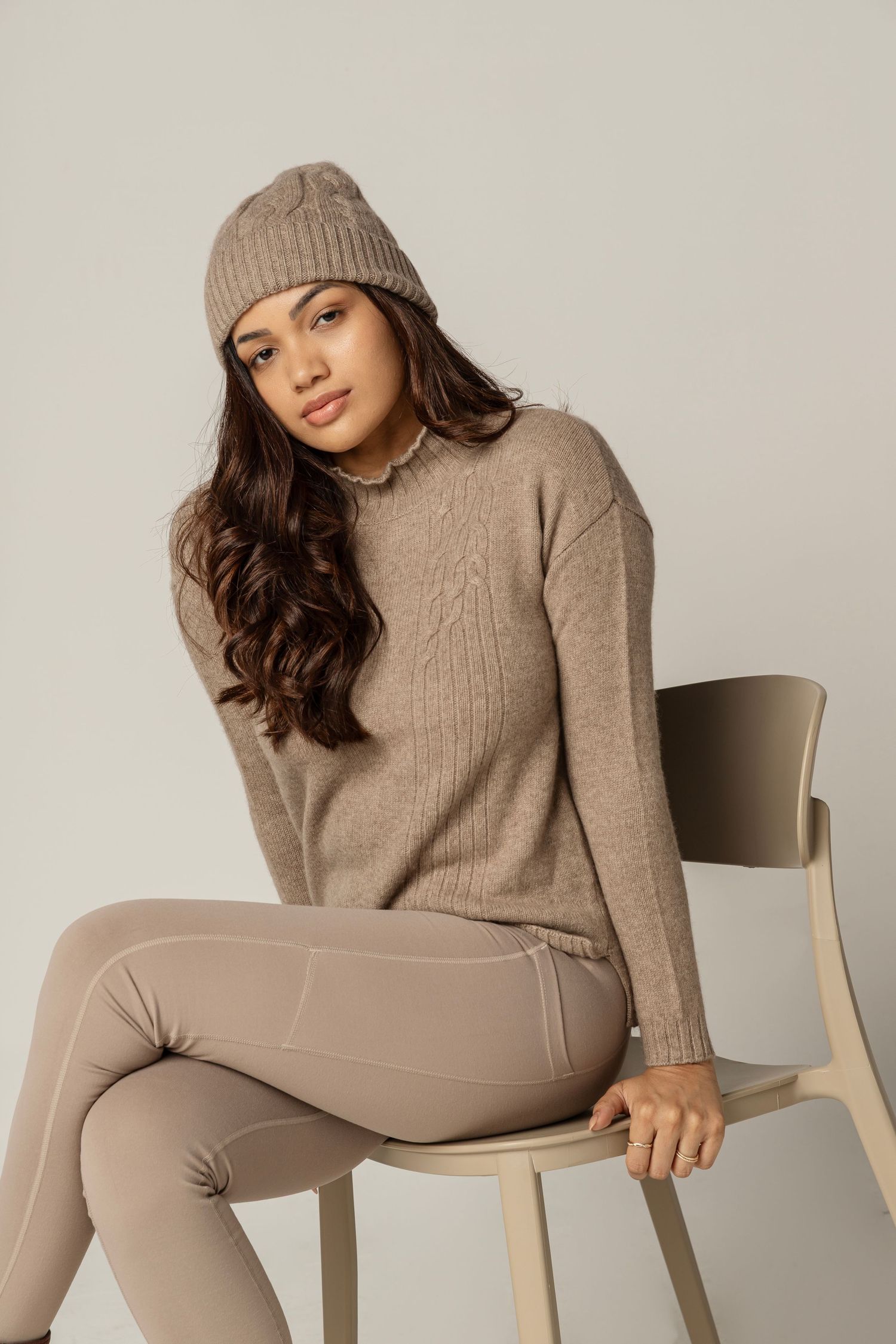 Fashion model wearing neutral beige loungewear and knit beanie poses on wooden chair in studio setting.