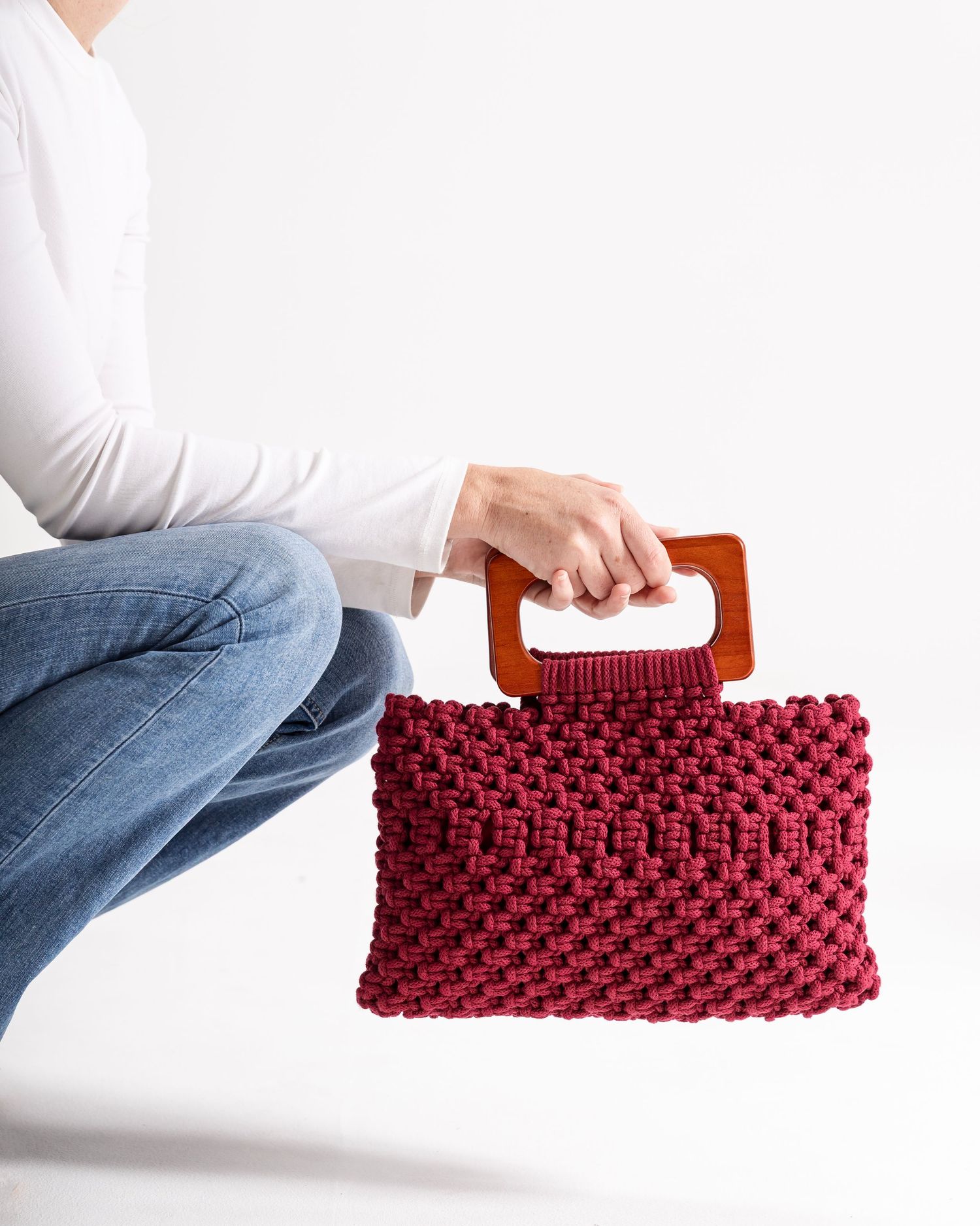 Person holding a burgundy macrame knit clutch bag with a wooden rectangular handle.