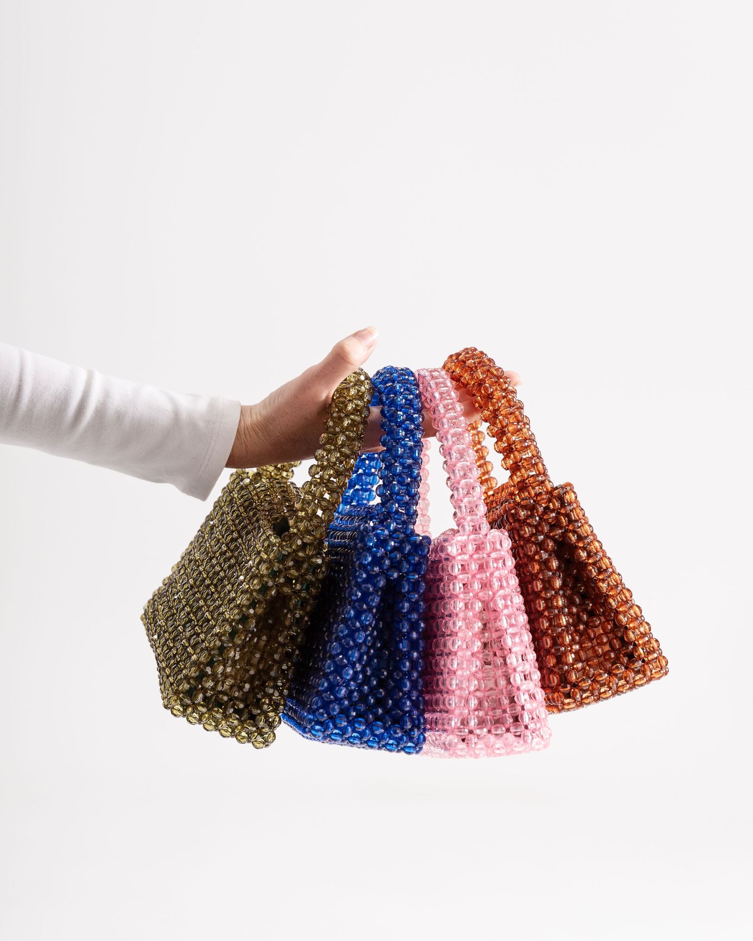 Hand holding four beaded handbags in olive, blue, pink, and brown colors against a white background.
