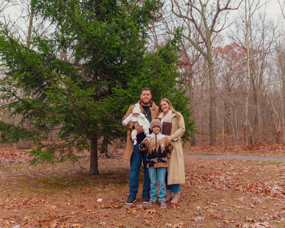 A family stands together outdoors in autumn, with fallen leaves on the ground and evergreen trees in the background.
