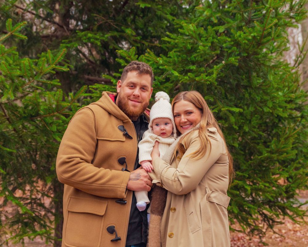 Three people in warm winter coats pose together outdoors against a backdrop of green evergreen trees.