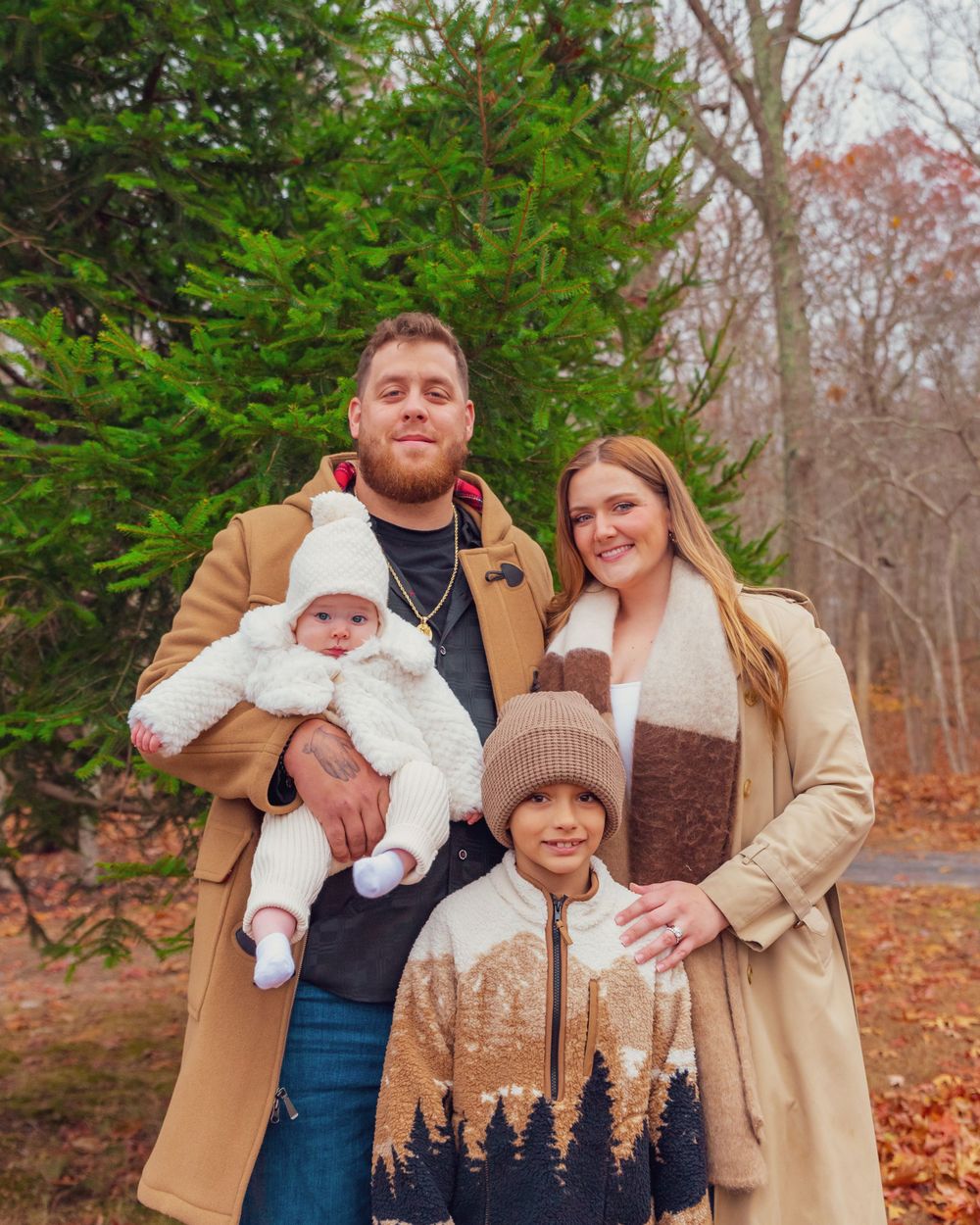 A family poses together for an autumn portrait in front of an evergreen tree with fallen leaves on the ground.