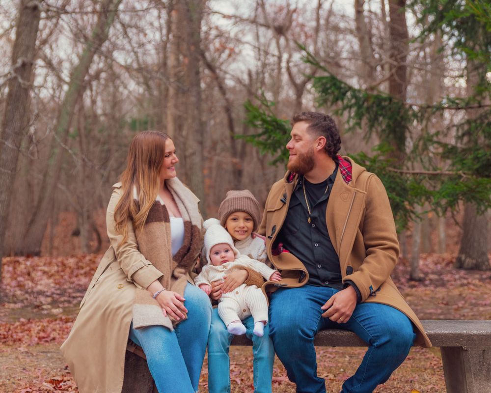 A family sitting together on a stone wall in autumn woods, wearing cozy coats and smiling at each other.