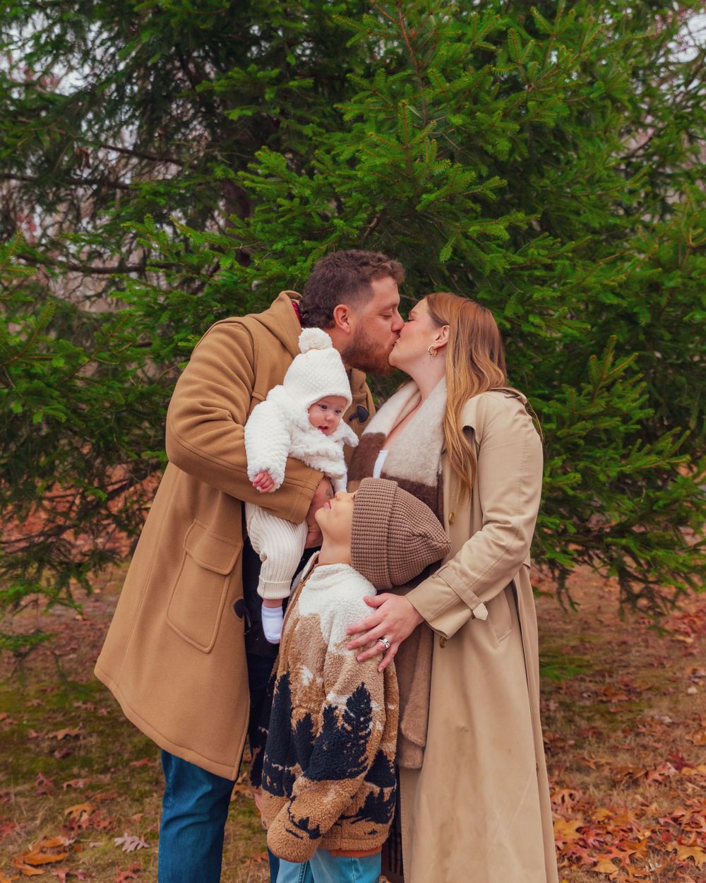 A family shares a tender moment together while wearing matching camel and beige coats in an autumn forest setting.