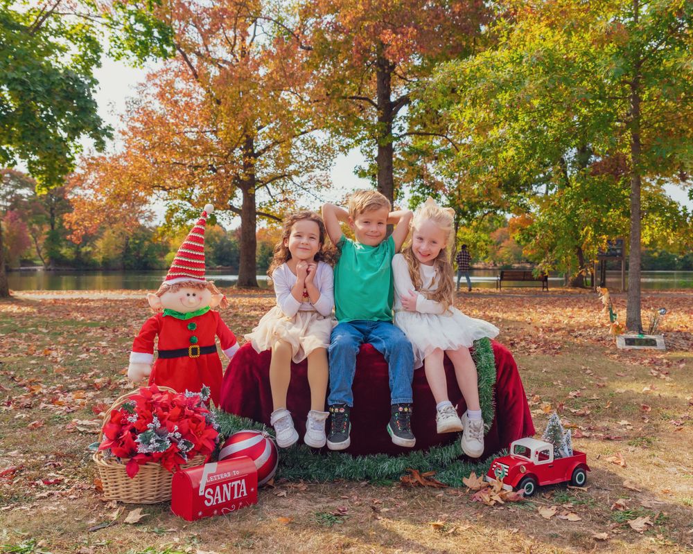 Four children in holiday outfits sit on red decorative sleigh surrounded by colorful autumn trees and falling leaves.