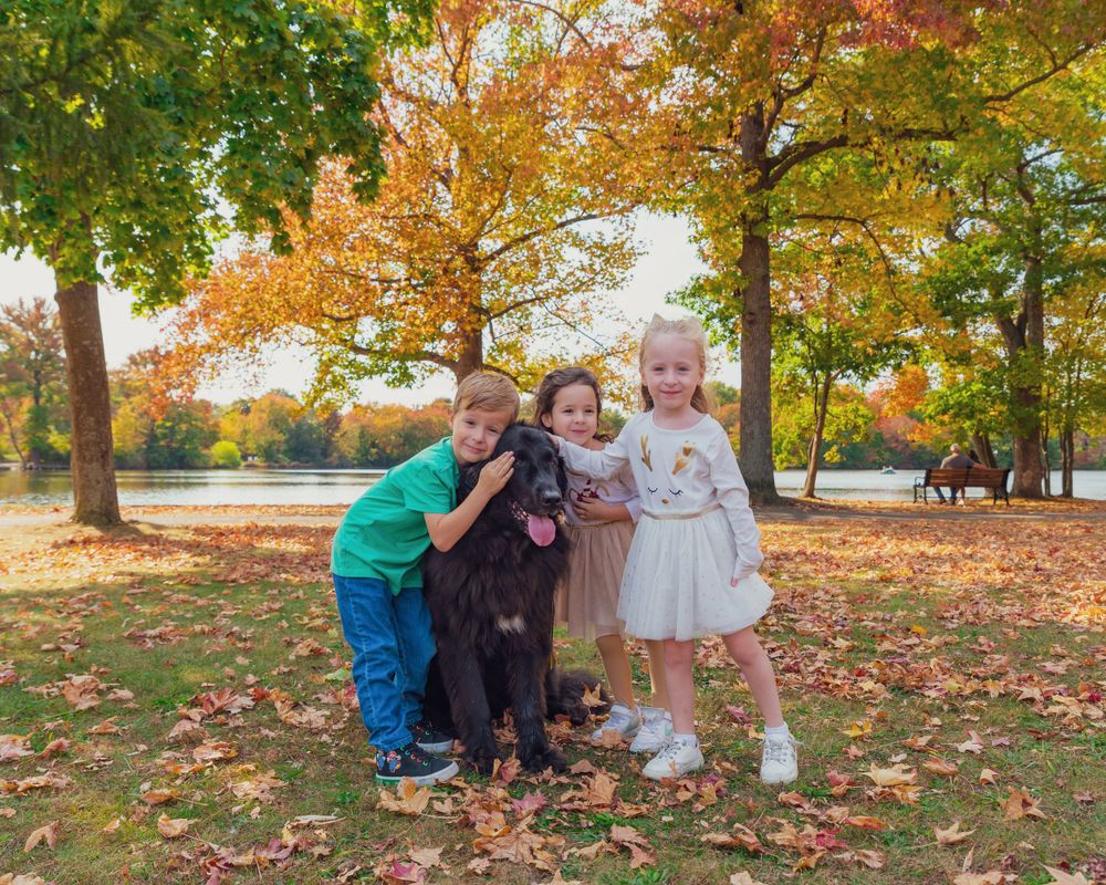 Children posing with black dog in autumn park with colorful fall foliage and lake in background.