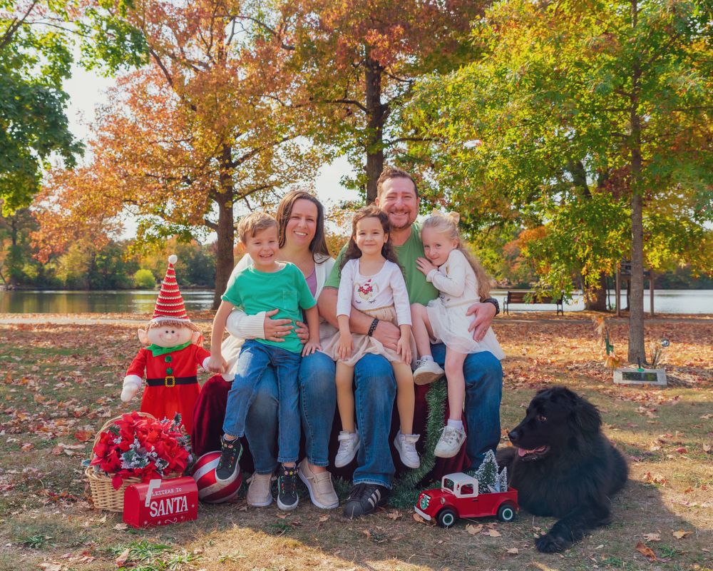 A group sits together outdoors in autumn with bright fall foliage, red wagons, and a black dog beside them.