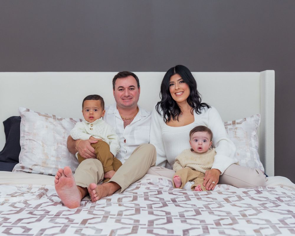 A happy family of four relaxes together on a white bed with a geometric patterned duvet cover.