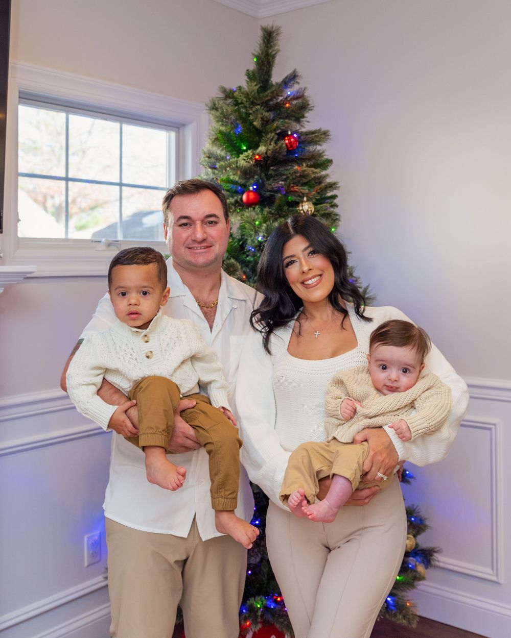 A family poses together in front of a decorated Christmas tree in a bright room with white wainscoting.